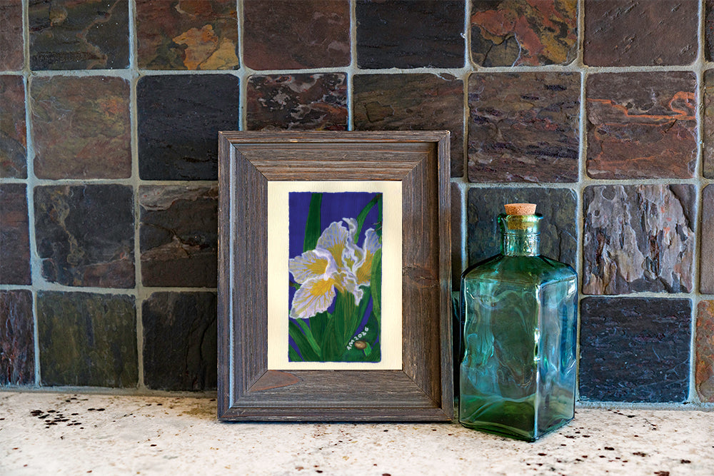 A framed art print depicting white irises on a dark blue background, styled on a stone surface next to a vintage green glass bottle against a dark slate tile wall.