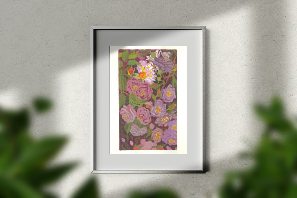 A framed floral art print featuring purple and pink flowers with green leaves, displayed on a textured light gray wall with soft shadows and blurred green foliage in the foreground.