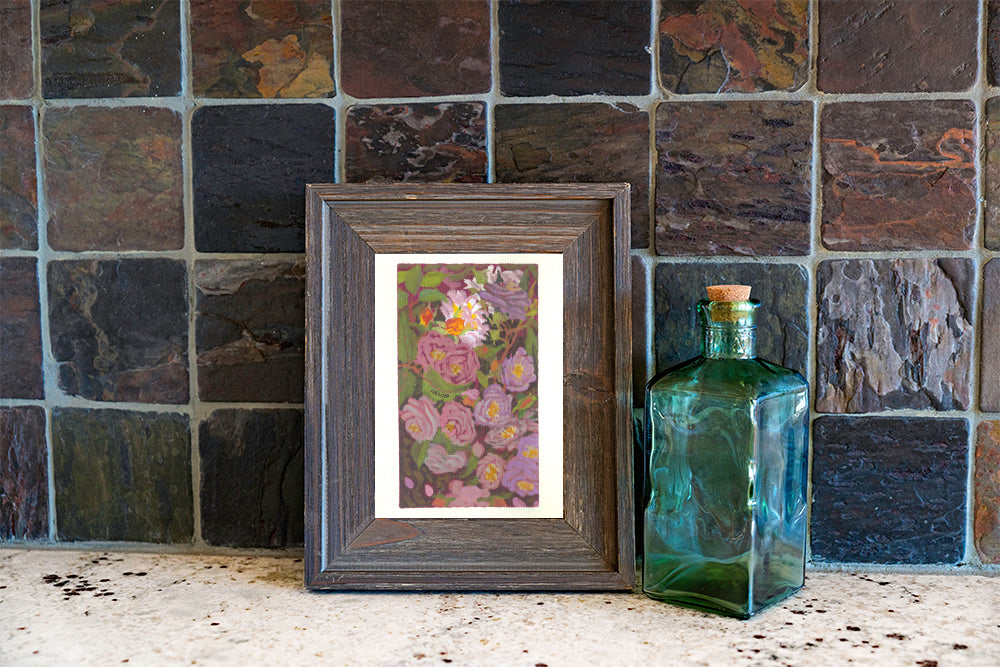 A framed floral art print with pink and purple flowers in a rustic dark wood frame, styled on a stone surface next to a vintage green glass bottle against a dark slate tile wall.